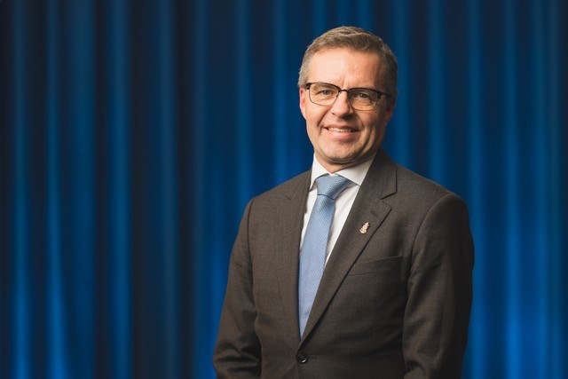 Magnus Wallhagen ler in i kameran i klädd kostym framför blå bakgrund.