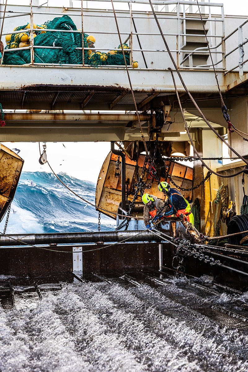 Two fishermen on a large fishing vessel haul in the net. Water splashes across the deck. In the background, high waves and breakers can be seen