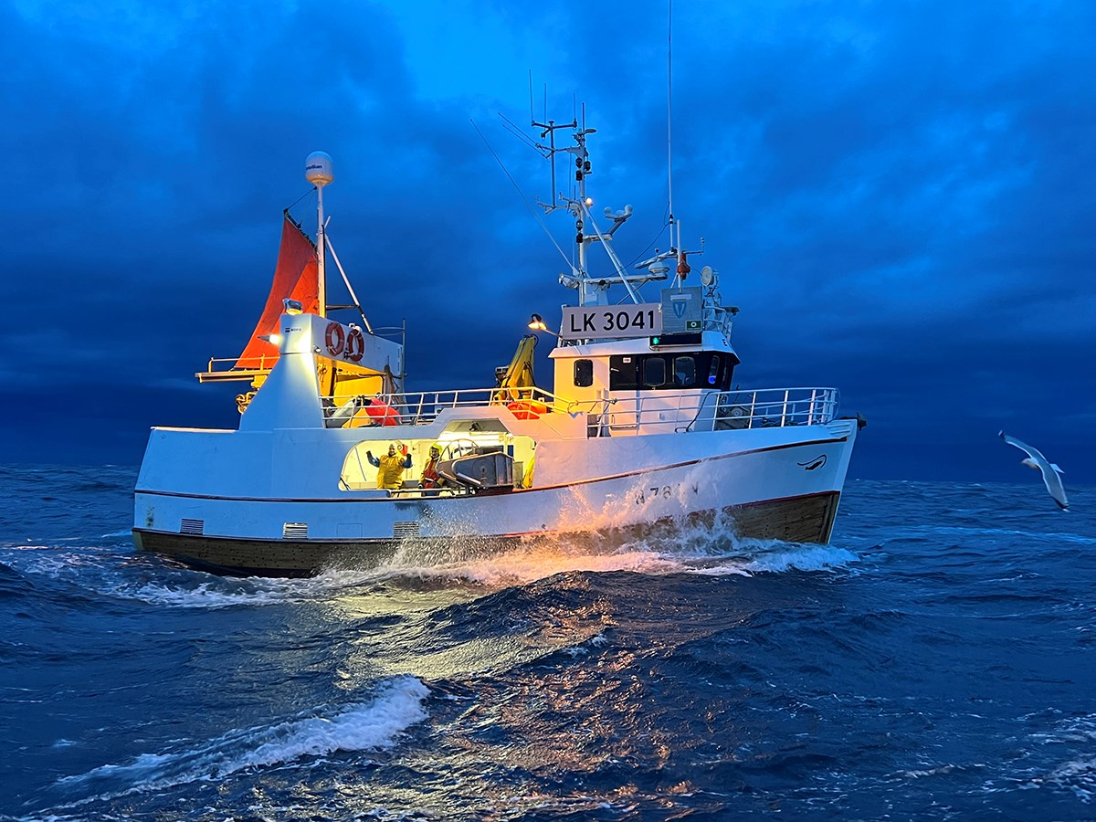 A fishing boat cuts through the waves. Two fishermen stand in an illuminated space, looking toward the camera. A seagull drifts in front of the boat. The background is storm‑like