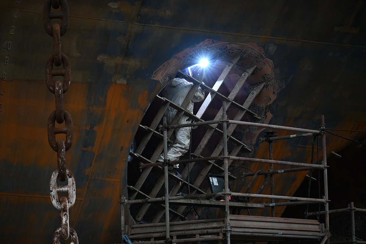 A section of a ship’s bow. Chafing marks are visible on the left, and a large anchor chain hangs down. A person in a white coverall clings to the large grating at the bow thruster while welding something inside.