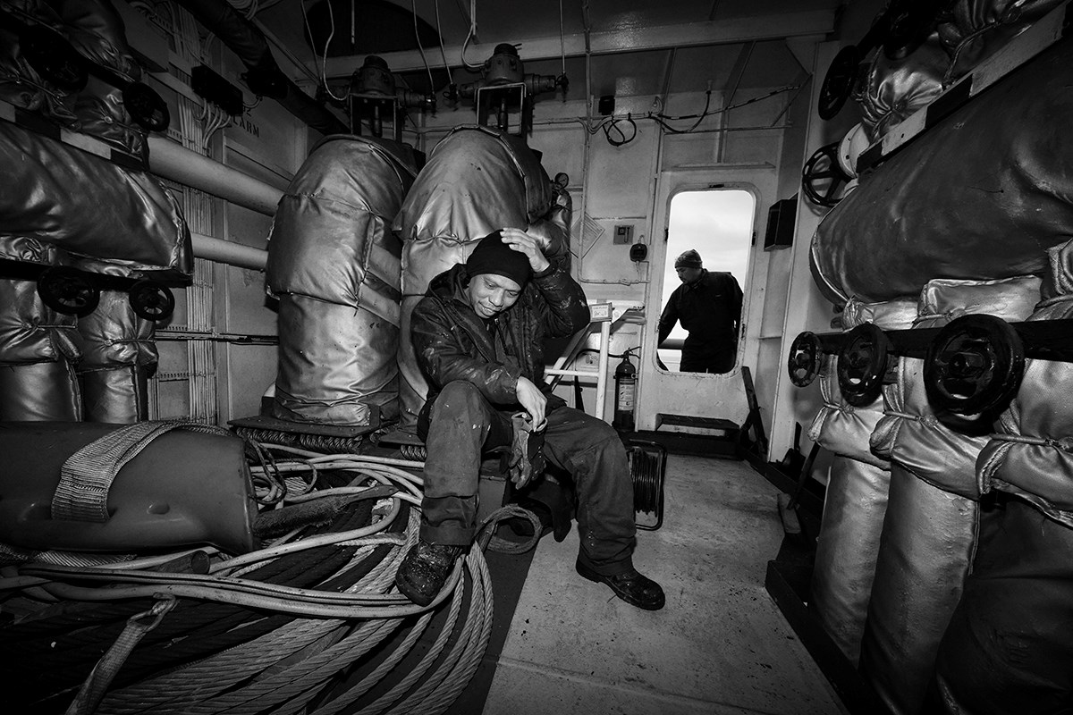 A black‑and‑white image of a man in a coverall sitting in a room with his foot resting on a thick cable, one hand on his head and the other holding a pair of work gloves. Through the window in the bulkhead (the door), another man can be seen working