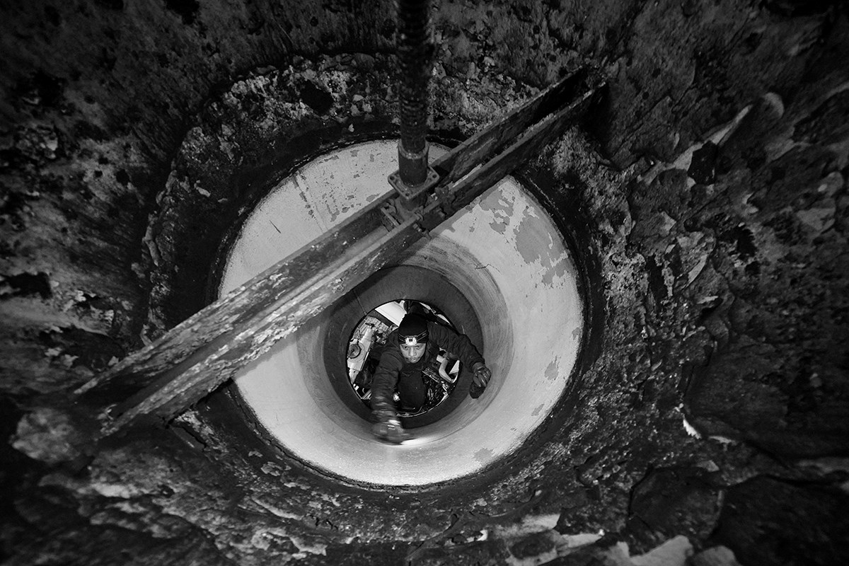 A black‑and‑white photo taken from above. A man wearing a headlamp can be seen working on something further down inside the pipe