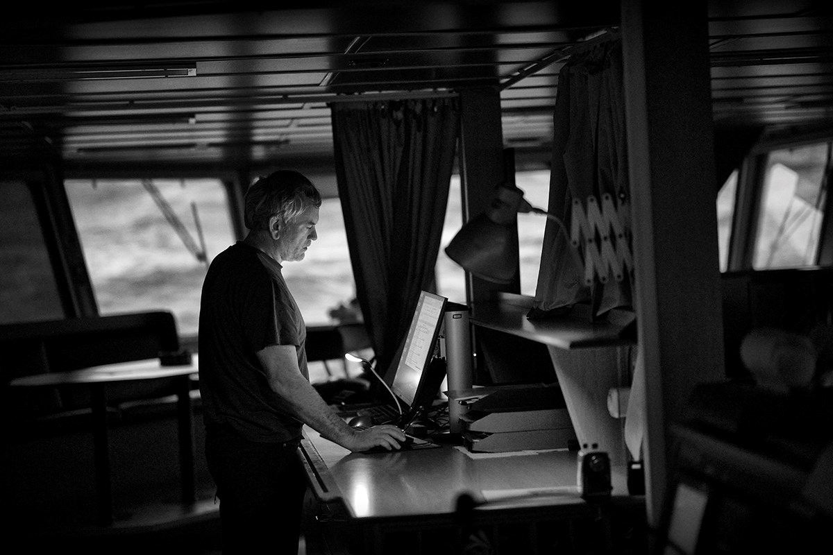A black‑and‑white photo of a man standing in front of a computer on the ship’s bridge. In the background, the sea can be seen through the bridge windows