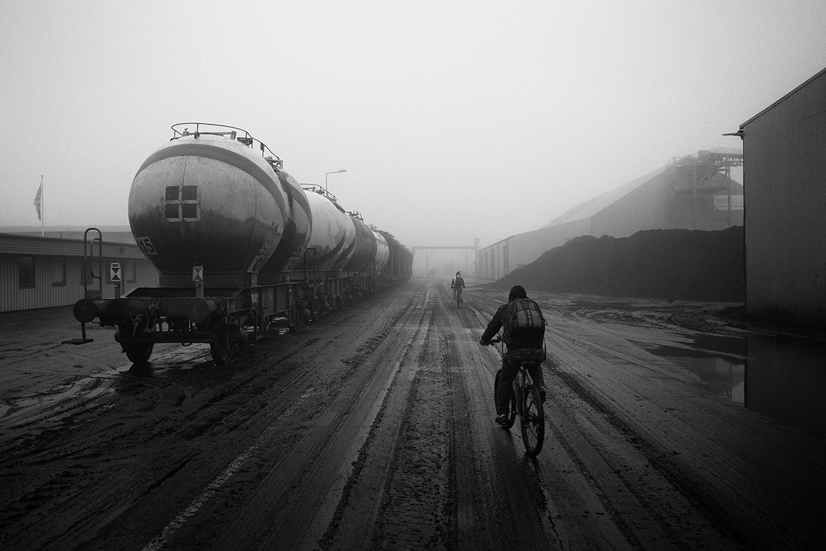 A black‑and‑white photo of two cyclists with backpacks riding toward the fog. To the left stands a train, and to the right there is a large pile of coal or a similar material