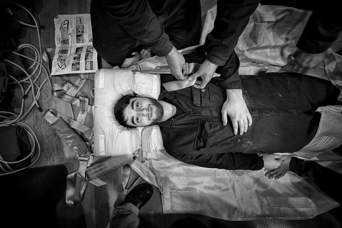 A black‑and‑white photo of a smiling man lying on a stretcher. His head is supported by a shaped cushion, and several other people’s arms and legs are visible in the frame