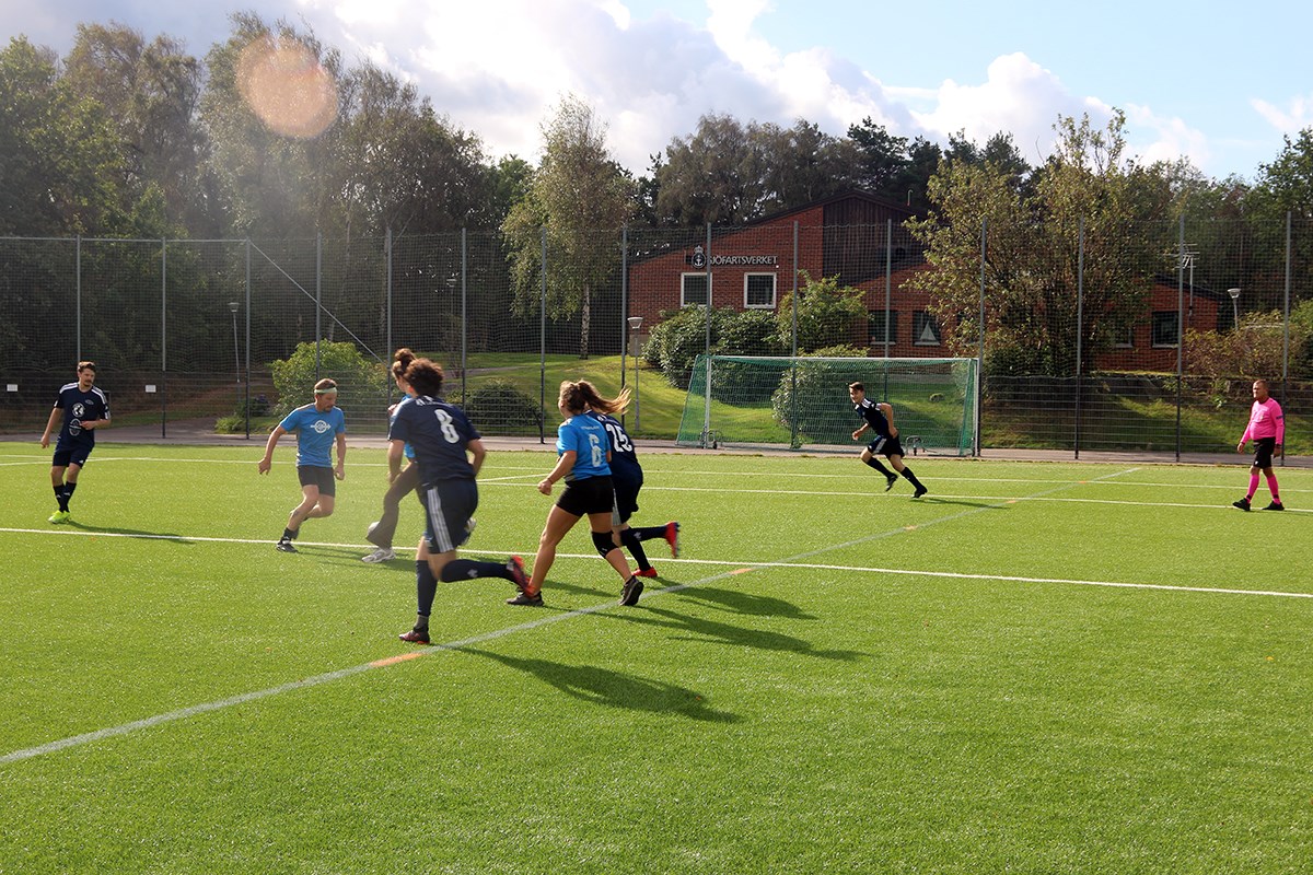 Flera fotbollsspelare springer, en gående domare i höger kant. Bakom ett fotbollsmål skymtar en tegelbyggnad med en Sjöfartsverket-skylt.