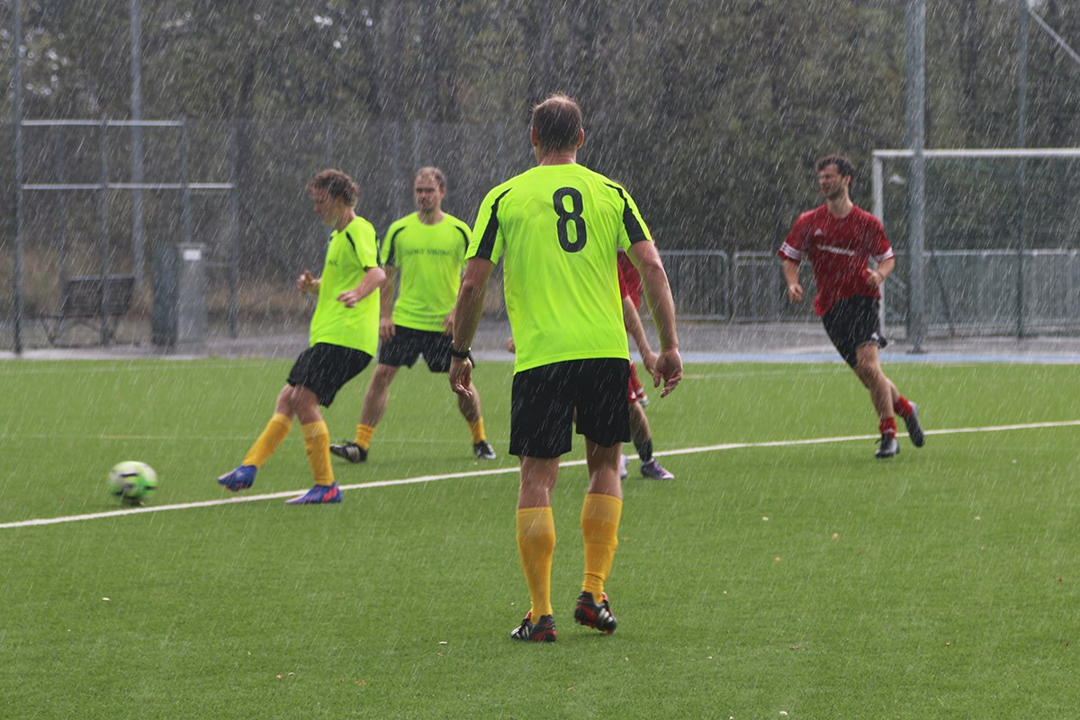 En spelare i neongrön dress (med ryggen mot kameran)  betraktar hur medspelarna passar bollen. Tre spelare i röda dressar syns också i bild. Allt medan regnet öser ned.