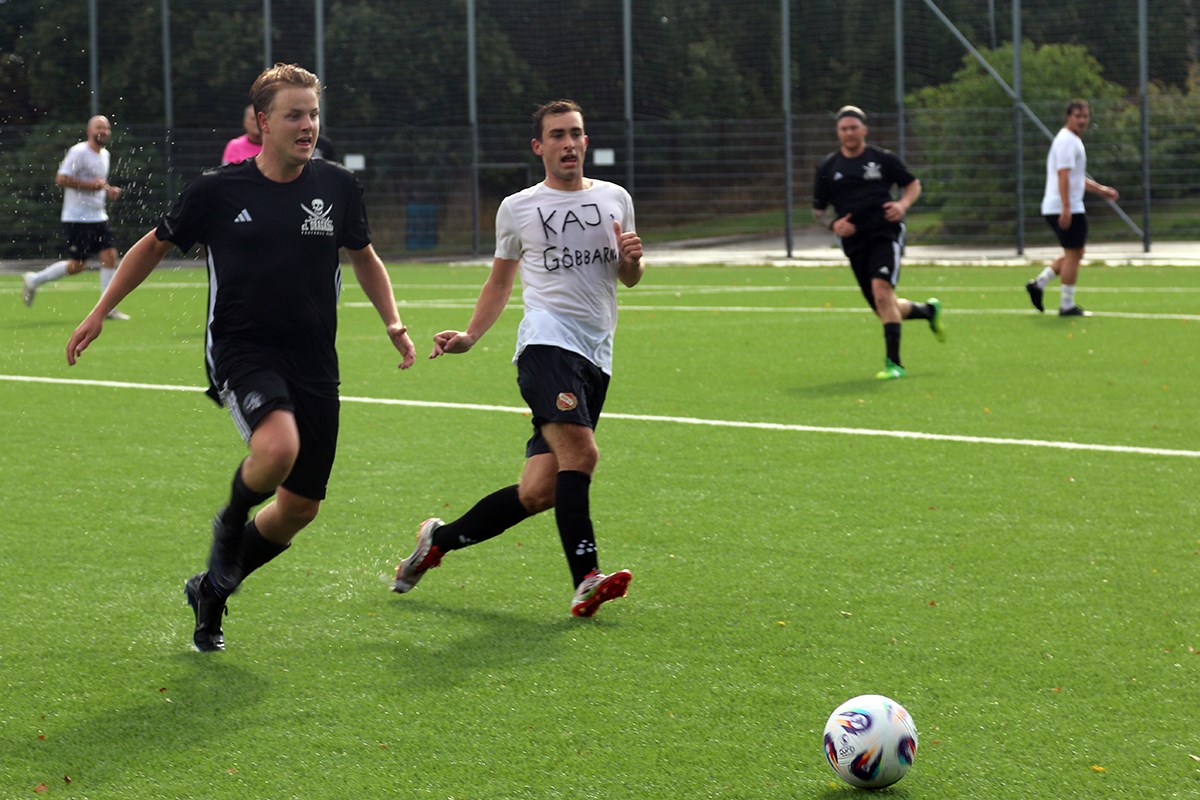 Två spelare jagar bollen, som rullar ned mot bildens högra hörn.  Vatten skvätter från både spelare och plan när de springer. I bakgrunden syns fler spelare.