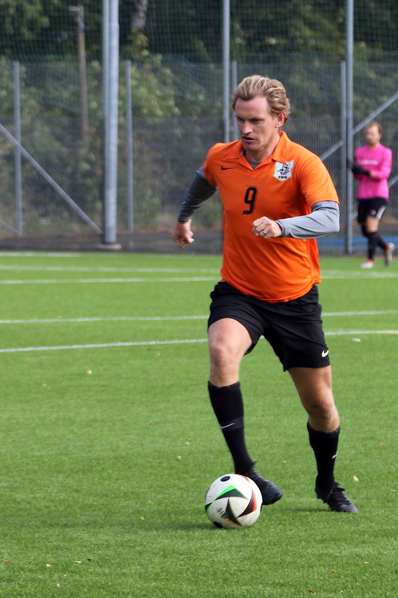 En ensam spelare driver bollen framåt. I bakgrunden skymtar en målvakt från en annan match på planen intill.