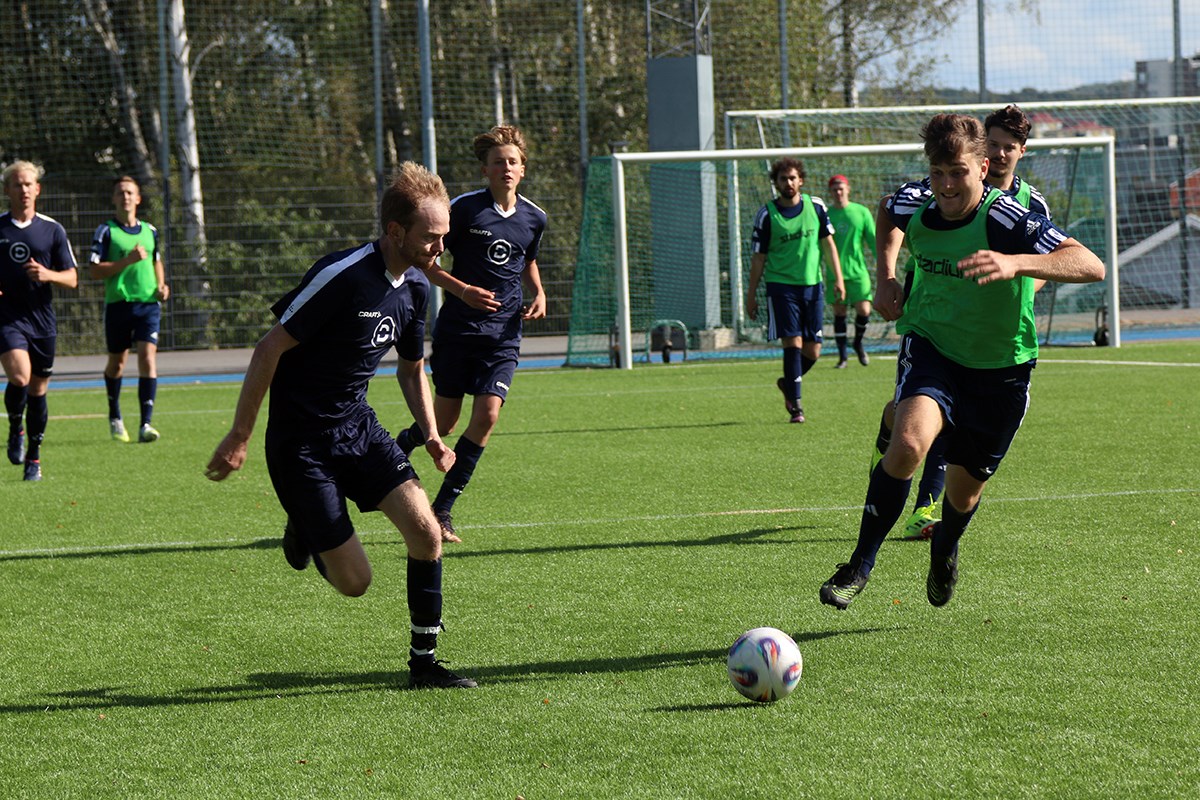 Två fotbollsspelare rusar mot bollen, en i blå dress och en i grön väst. I bakgrunden syns fler spelare.