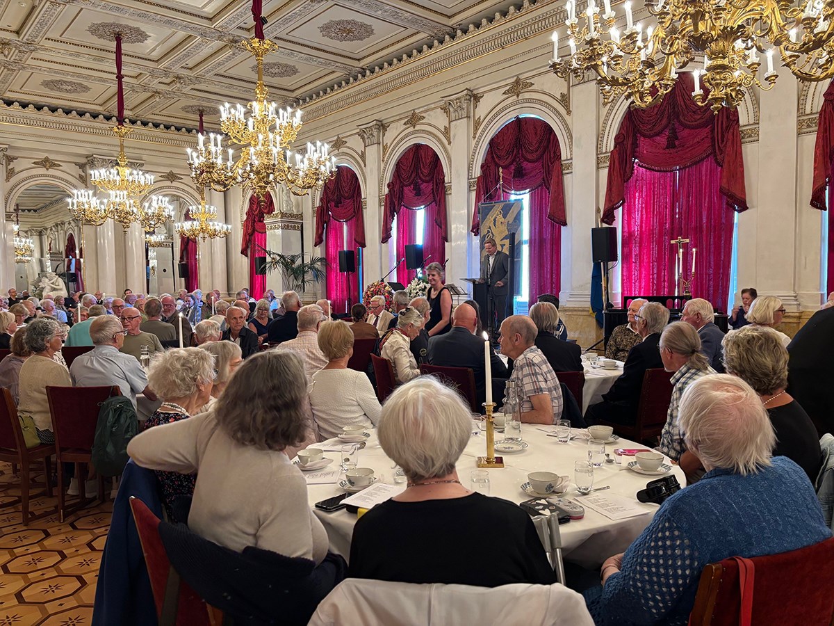 Många personer sitter vid flera runda bord, i vacker salong, och tittar mot scenen, där en talare står. Kristallkronor i taket.