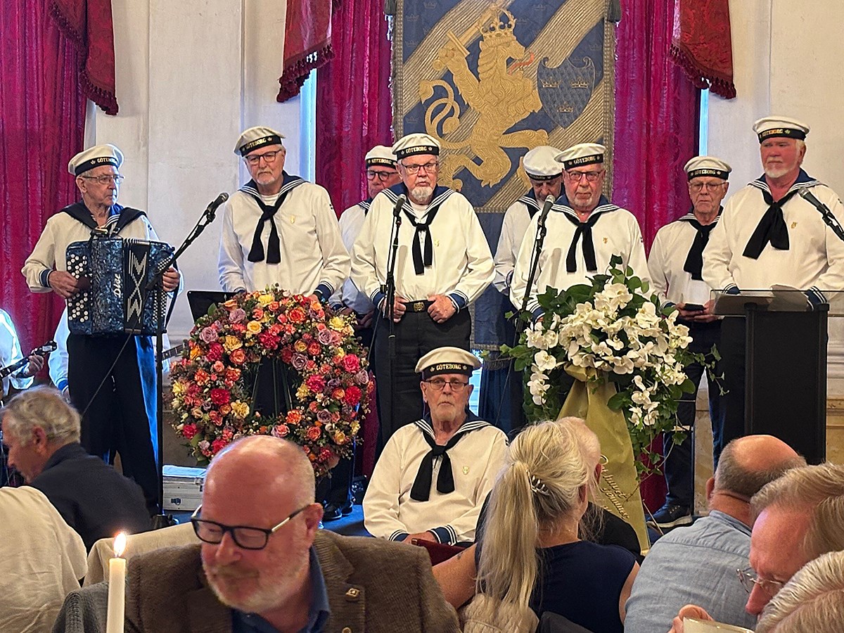 Åtta personer i sjömanskostymer och -mössor står på scenen och underhåller. I förgrunden ses några av åhörarna fika