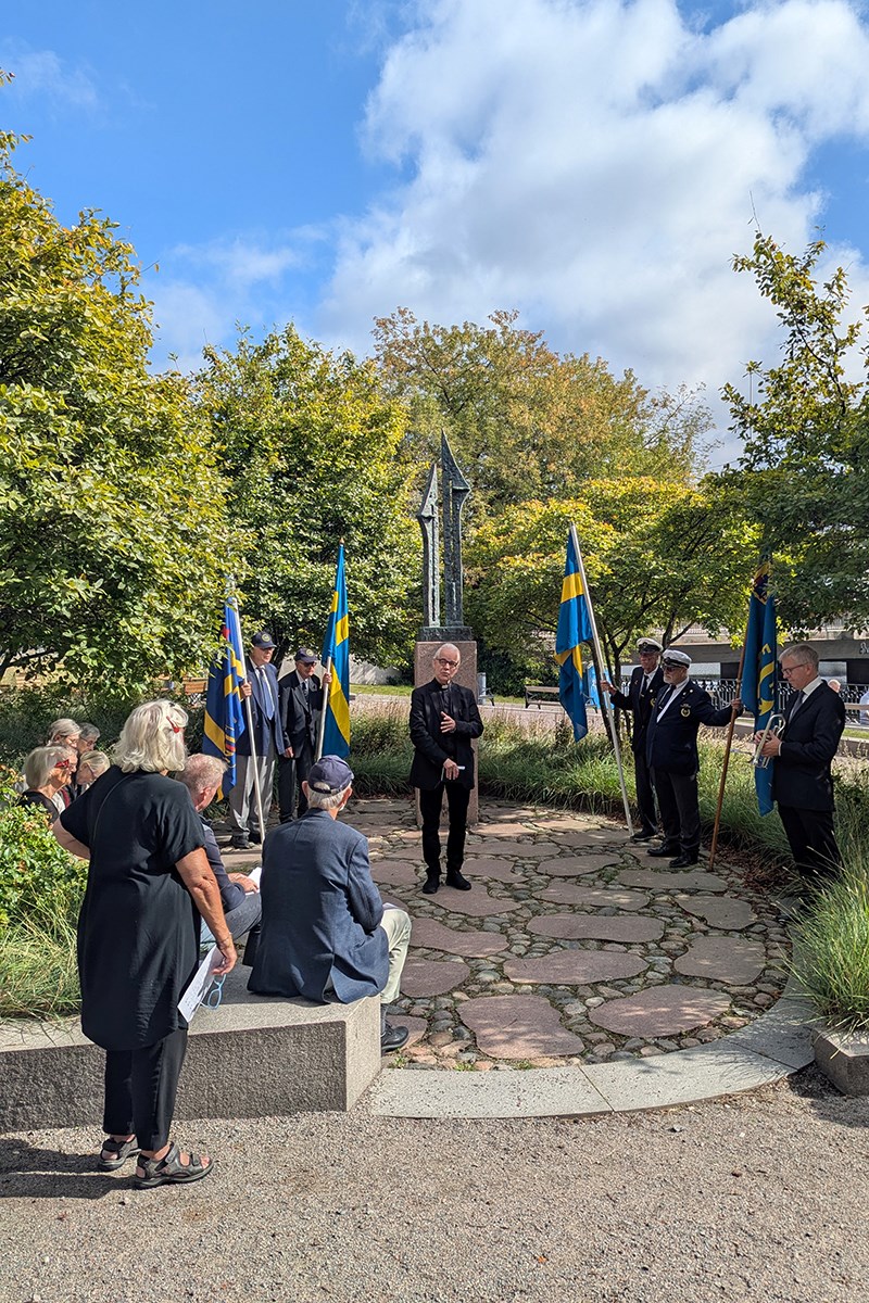 En grupp lyssnar till en präst som står och håller ett korum, vid ett minnesmärke. Några av åhörarna håller i flaggor.